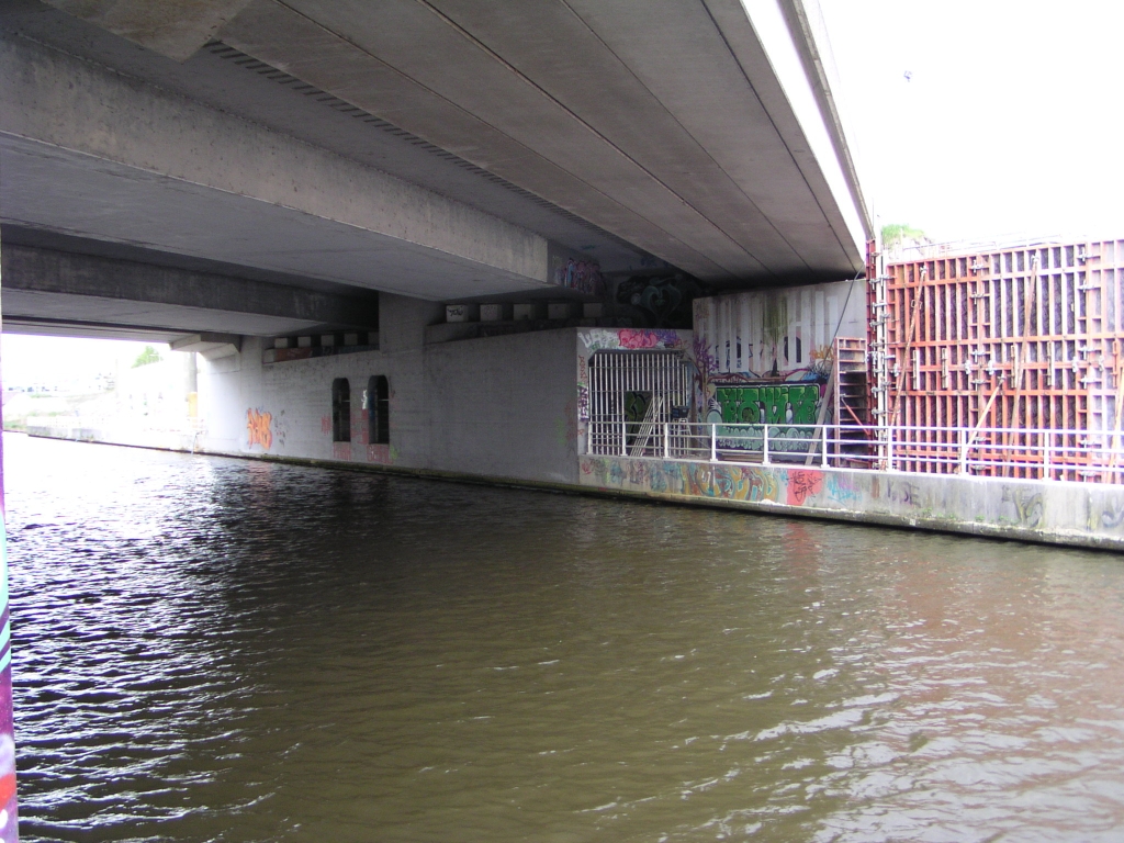 p4300031.jpg - Zo ziet zo'n tunneltje eruit, thans gebarricadeerd met tralies. Er moet veel te vinden zijn over de historie van deze bruggen, maar niet op internet. Dan maar wat observeren en gokken... op een topografische kaart uit 1986 zien we de aansluiting Veldhoven nog slechts gerealiseerd als een halve aansluiting, met korte in- en uitvoegers in noordelijke richting - naar deze brug toe. Vermoedelijk was toen alleen het middelste deel van de brug aanwezig. De in/uitvoegstroken liepen dus niet door over de brug, of men had de vluchtstroken moeten opgeven. Later, misschien tijdens het project tot completering van de aansluiting Veldhoven, is de brug verbreed, getuige de sterk afwijkende bouwstijl van het rechtergedeelte in de foto. Dat bestaat uit prefab liggers, terwijl het oude middendeel in situ gestort lijkt te zijn. Misschien werden de tunneltjes ooit als eerste gebouwd (ca. 1968) en gebruikt als steunen voor bekistingsdragers over het water heen. Interessant is ook het verschil in landhoofdconstructie tussen de twee verbredingen. Aan de linker (oost-) kant is het tunneltje verlengd om er de liggers op te kunnen leggen, analoog aan de werkwijze met het oude middendeel. Aan de rechter (west-) zijde daarentegen rusten de liggers op een eigen landhoofd. Aan de zuidzijde van de brug (standpositie) is de situatie precies omgekeerd.